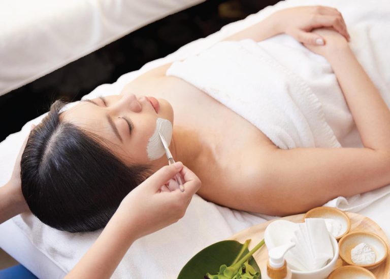 Hands Of Spa Therapist Applies Facial Product To A Woman Laying On Massage Table
